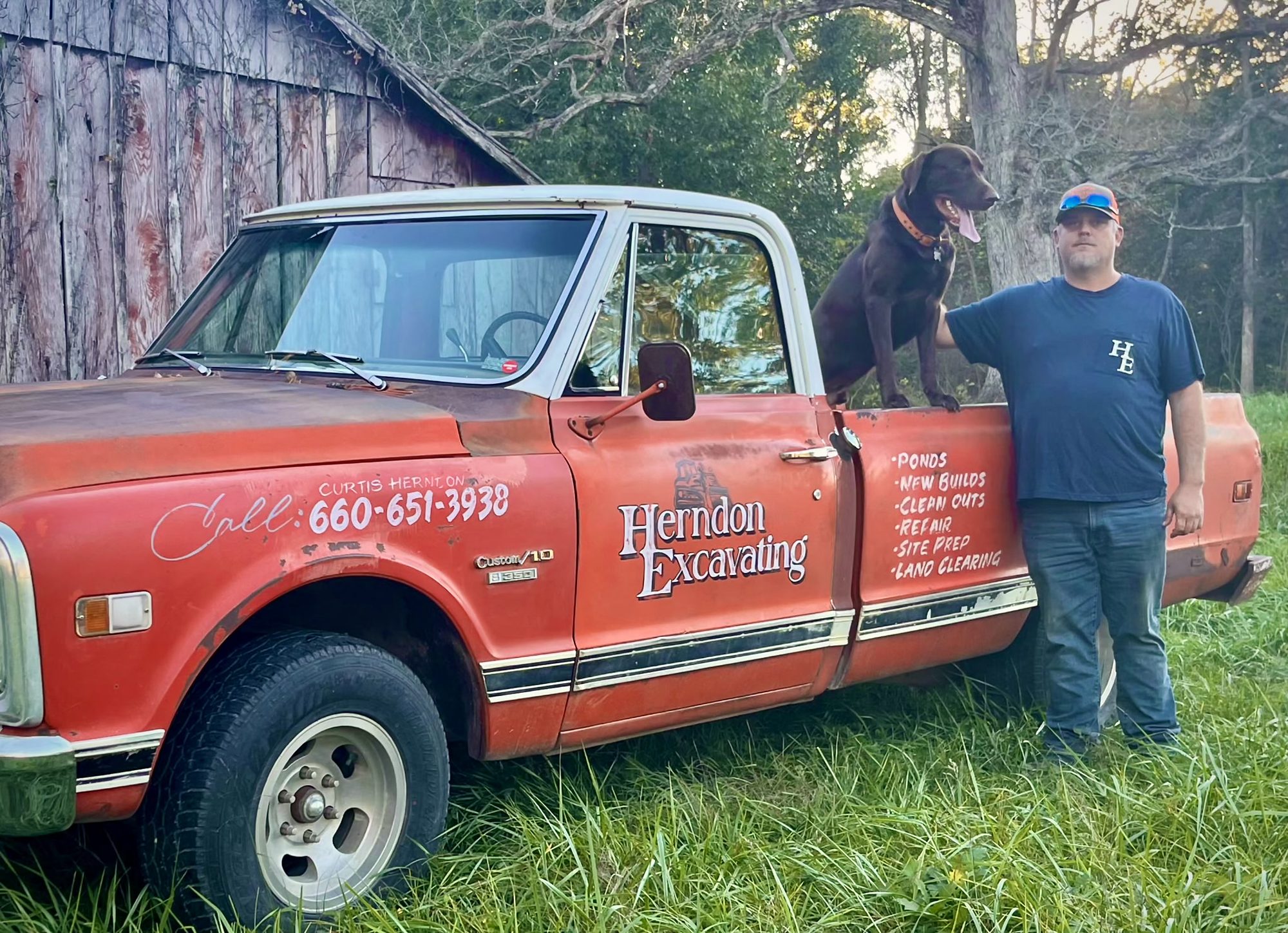 Curtis Herndon and his dog beside the Herndon Excavating pickup, a hand-lettered red Chevrolet C10, in front of a weathered barn